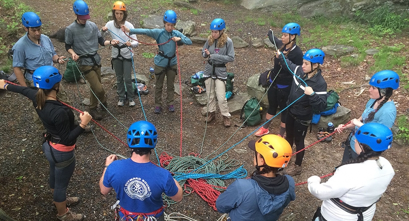 a group of people wearing helmets stand in a circle and are connected by a tangle of ropes at the center. 